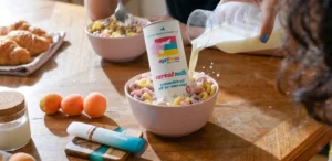 A cropped, closer view of the breakfast scene showing milk pouring into a bowl of cereal around an upright Ayrloom packaging tube, next to three apricots and a disposable vape pen on a coaster.
