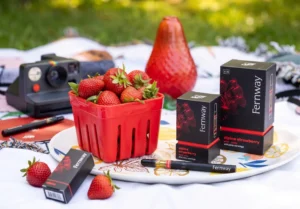 A bright, outdoor lifestyle product photograph showcasing 'Fernway' cannabis vape products arranged in a scenic picnic setting. In the center, a decorative white serving tray holds a classic red plastic berry basket overflowing with fresh, ripe strawberries. Arranged on the tray are sleek black packaging boxes with red text branding for Fernway's 'alpine strawberry' wild juice cartridge. A slim, black Fernway vape pen with a clear oil reservoir filled with golden distillate lies horizontally in the foreground. In the softly blurred background, a vintage black instant camera and a red glass vase rest on a white blanket spread over a green grassy lawn, emphasizing a fresh, fruit-flavored summer theme.