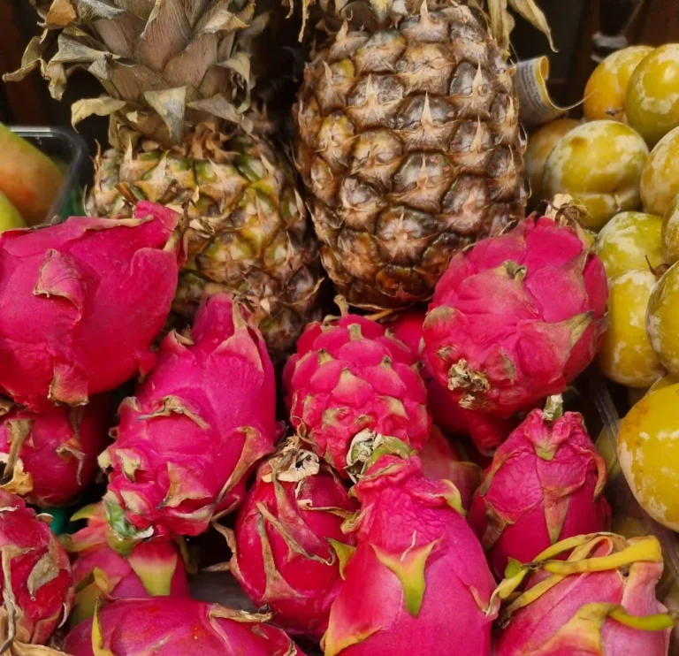 A vibrant, close-up photograph featuring a dense arrangement of fresh exotic fruits. In the foreground, a prominent cluster of bright magenta dragon fruits (pitayas) showcases their characteristic green-tipped scales. Nestled directly behind them are two textured, rough-skinned pineapples with sharp, spiky crowns. To the right, a pile of smooth, round yellow-green fruits adds to the variety. The rich, contrasting colors and textures create a lush, appetizing display that vividly evokes a sweet, refreshing, and bold tropical flavor profile.